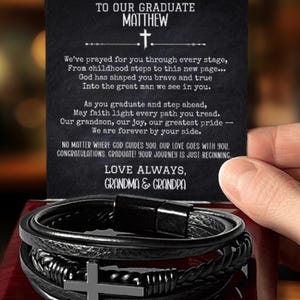 May include: A black leather bracelet with a silver cross charm, presented in a dark wooden box. A card with the text "TO OUR GRADUATE MATTHEW" and a heartfelt message is held above the bracelet.