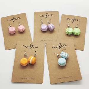 Könnte beinhalten: Fünf Paare Macaron-Ohrringe auf braunem Karton mit dem Wort "craftit" und der Website "www.craftitshop.com" auf jeder Karte gedruckt. Die Macarons sind in den Farben Lavendel, Grün, Hellblau, Orange und Rosa.