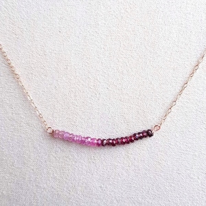 May include: A delicate gold chain necklace with a graduated row of pink and red gemstones. The necklace features a subtle ombre effect, transitioning from light pink to deep red.