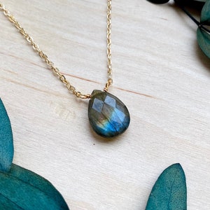 May include: A gold chain necklace with a teardrop-shaped labradorite gemstone pendant. The pendant has a dark blue and green iridescent sheen.