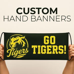 May include: A black hand banner with the text "CUSTOM HAND BANNERS" at the top. The banner features a yellow tiger logo and the words "GO TIGERS!" in yellow. The banner is held up by two hands.