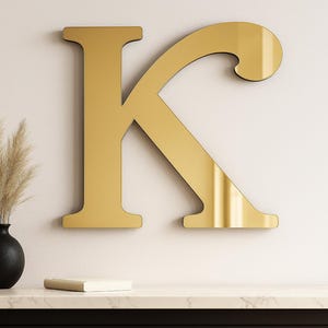 May include: A large, gold-colored letter "K" mounted on a wall. Below, a black vase holds dried ornamental grass. A small, closed book rests on a marble-topped table. The letter is approximately 30 inches tall.