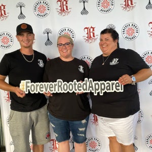 Pu&ograve; includere: Tre persone indossano magliette nere con un logo bianco che dice "Forever Rooted Apparel" e un disegno di nodo celtico. Le persone tengono un cartello che dice "#ForeverRootedApparel".