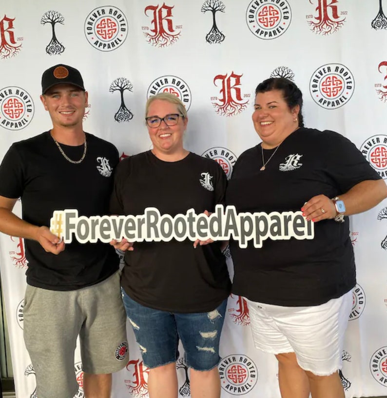 Pu&ograve; includere: Tre persone indossano magliette nere con un logo bianco che dice "Forever Rooted Apparel" e un disegno di nodo celtico. Le persone tengono un cartello che dice "#ForeverRootedApparel".