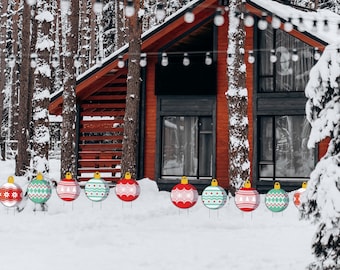 Puede incluir: Una escena invernal con una cabaña roja, luces de cadena y árboles cubiertos de nieve. En primer plano, decoraciones de jardín de adornos navideños coloridos con varios patrones se exhiben en la nieve, agregando un toque festivo.