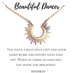 May include: Gold necklace with a sunburst pendant. The necklace has a message that reads "You have a beautiful gift and your hard work and effort have paid off. When it comes to dancers, you shine the brightest." The message is signed "ROCO & JO".