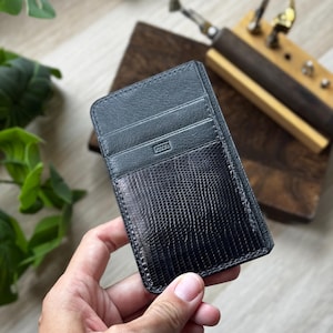 Può includere: Un porta carte in pelle nera con un motivo strutturato a forma di rettile sul davanti. Il porta carte ha più fessure e una tasca con cerniera. Il nome del marchio è impresso sul davanti. Il porta carte è tenuto in mano.