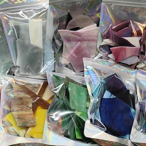 May include: Multiple clear, iridescent, resealable plastic pouches filled with assorted shards of stained glass in various colors including white, grey, pink, purple, green, blue, yellow, and brown, showcasing diverse textures and opacities.