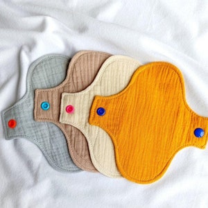 May include: Four reusable cloth menstrual pads in different colors: gray, beige, light brown, and yellow. Each pad has a blue snap button on one side and a colored snap button on the other side.