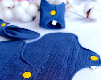 Puede incluir: Un juego de tres compresas menstruales de tela reutilizables azules con broches amarillos. Las compresas están etiquetadas con la talla "M".