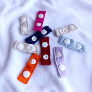 May include: A selection of fabric button extenders in shades of pink, navy, red, orange, and beige. Each extender features two white buttons. The extenders are displayed on a white, textured surface.
