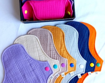 Puede incluir: Conjunto de siete compresas menstruales de tela reutilizables en varios colores, incluyendo morado, beige, naranja, gris, azul y naranja. Cada compresa tiene un cierre a presión y está etiquetada con una talla "M".