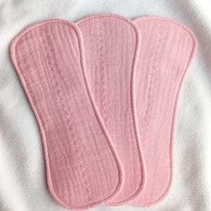 May include: Three pink, oblong cloth pads with a zig-zag stitch down the center. The pads are made of a textured fabric and have rounded edges. They are arranged on a white, textured surface.