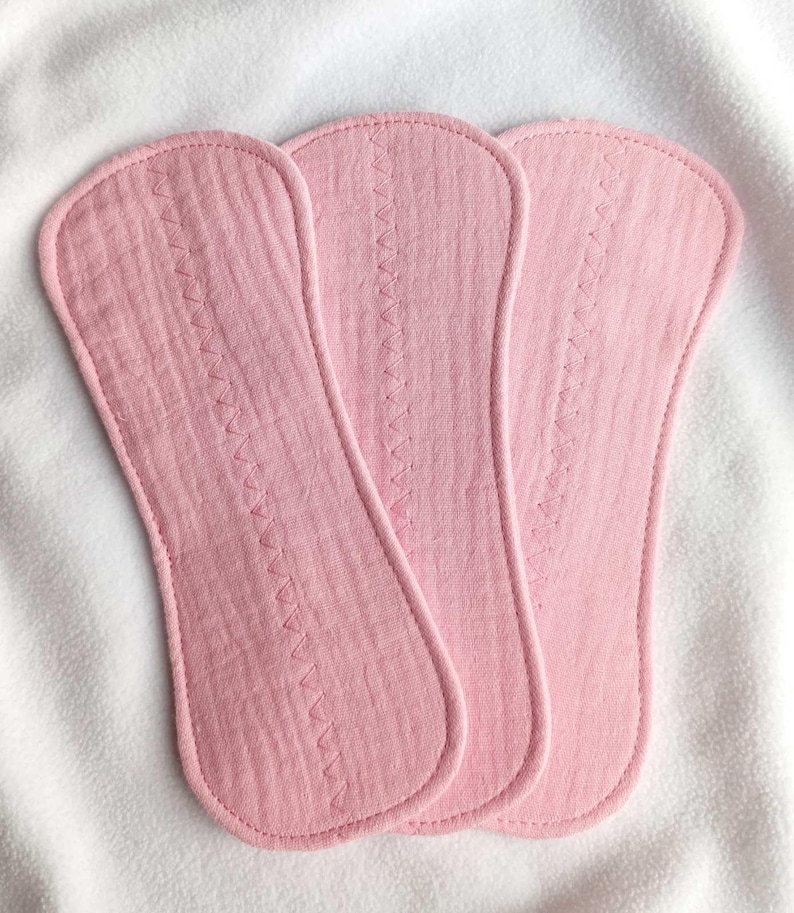 May include: Three pink, oblong cloth pads with a zig-zag stitch down the center. The pads are made of a textured fabric and have rounded edges. They are arranged on a white, textured surface.