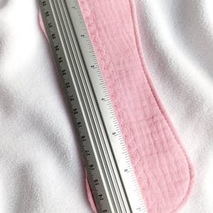 May include: A light pink reusable menstrual pad with a stitched edge, measuring approximately 11 inches in length. A metal ruler is placed next to the pad for measurement, set against a white background.