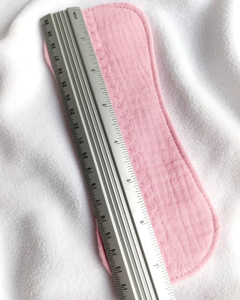 May include: A light pink reusable menstrual pad with a stitched edge, measuring approximately 11 inches in length. A metal ruler is placed next to the pad for measurement, set against a white background.