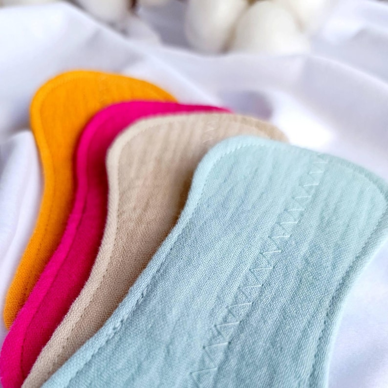 May include: Close-up of four reusable cloth menstrual pads in vibrant colors: orange, hot pink, beige, and light blue. The pads are arranged in a diagonal row, showcasing their soft, textured fabric and stitched edges. The background is a white, textured fabric.