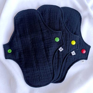 Puede incluir: Tres compresas menstruales de tela reutilizables de color azul oscuro con cierres de botón a presión de colores. Cada compresa tiene un botón a presión de diferente color: verde, amarillo y rojo. Las compresas están dispuestas sobre una superficie blanca. Cada compresa tiene una pequeña etiqueta blanca con la letra "M".
