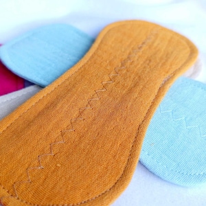 May include: Close-up of a mustard-colored, oblong reusable cloth menstrual pad with a zig-zag stitch down the center. Other colorful pads are visible in the background, including blue, pink, and gray.