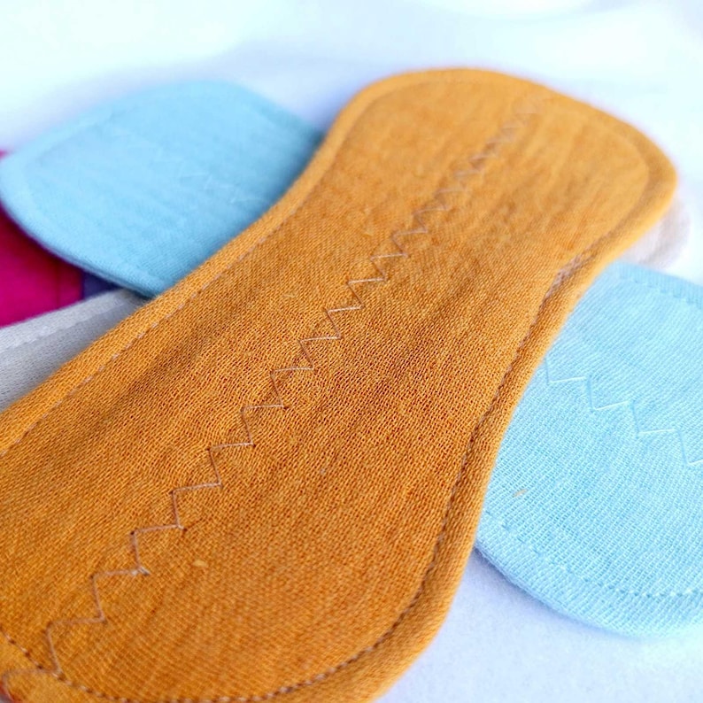 May include: Close-up of a mustard-colored, oblong reusable cloth menstrual pad with a zig-zag stitch down the center. Other colorful pads are visible in the background, including blue, pink, and gray.