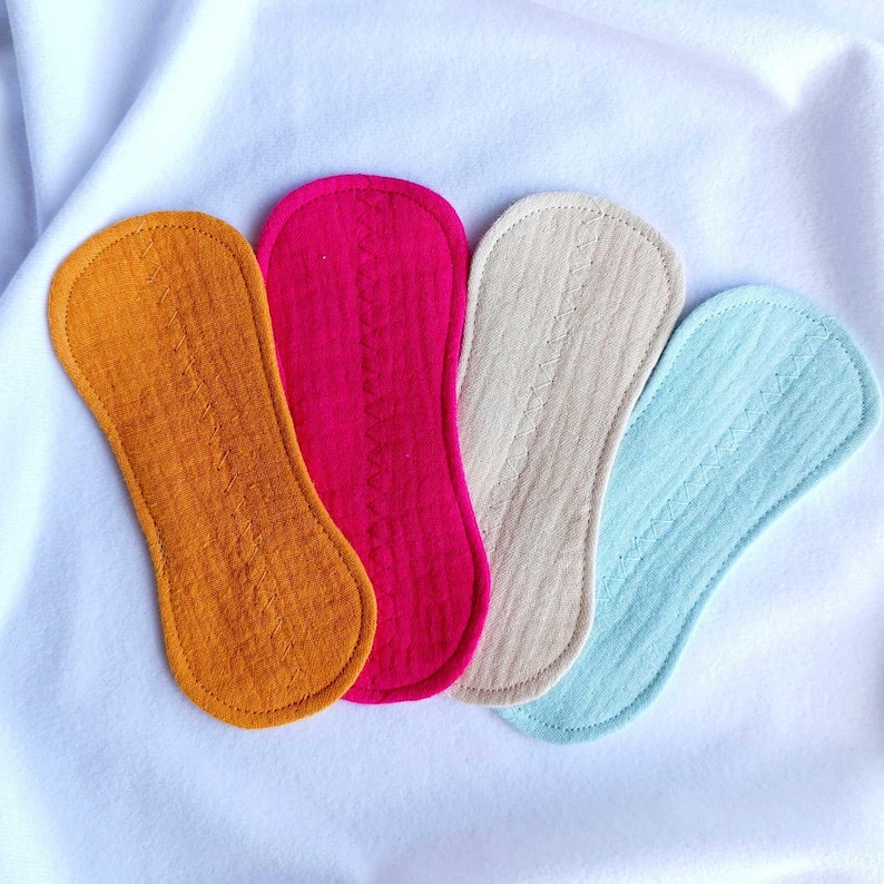 May include: Four reusable cloth menstrual pads in shades of orange, fuchsia, beige, and light blue. Each pad has a rounded shape and is made of textured fabric. The pads are arranged on a white surface.