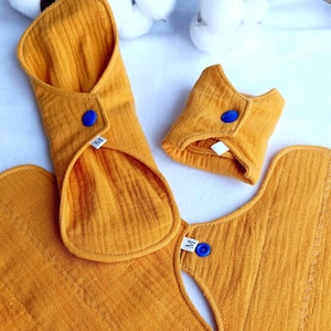 May include: Three reusable cloth menstrual pads in a mustard yellow color. The pads are folded and have a blue snap button on each. The pads are labeled with the letter "M" for size.