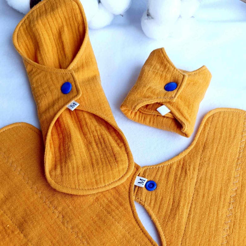 May include: Three reusable cloth menstrual pads in a mustard yellow color. The pads are folded and have a blue snap button on each. The pads are labeled with the letter "M" for size.