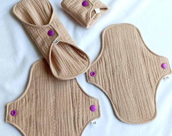 Puede incluir: Tres compresas menstruales de tela reutilizables en color beige con broches de presión morados. Las compresas son de diferentes tamaños y formas. La compresa más grande tiene forma de lágrima y tiene una etiqueta que dice "M".