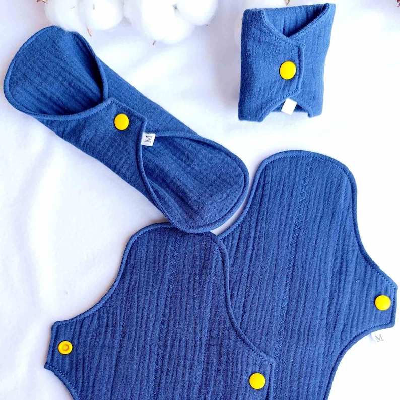 May include: A set of three blue reusable cloth menstrual pads with yellow snaps. The pads are in different sizes, with the largest pad having the letter "M" printed on it.