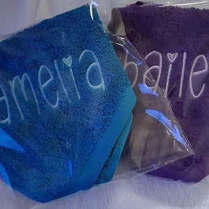 May include: Two plush towels individually wrapped in clear plastic. One towel is bright blue with the name amelia embroidered in light blue thread. The other towel is purple with the name bailey embroidered in white thread. Both feature a heart icon.