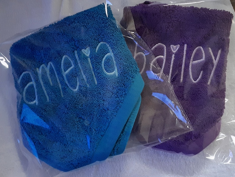 May include: Two plush towels individually wrapped in clear plastic. One towel is bright blue with the name amelia embroidered in light blue thread. The other towel is purple with the name bailey embroidered in white thread. Both feature a heart icon.