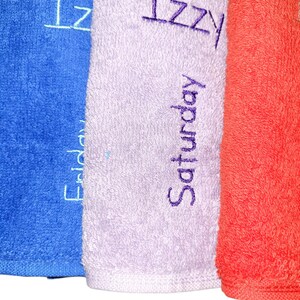 May include: Three folded terry cloth towels in blue, light purple, and orange. The blue towel features the embroidered name Izzy and the word Friday. The light purple towel displays the name Izzy and the word Saturday in dark purple thread.