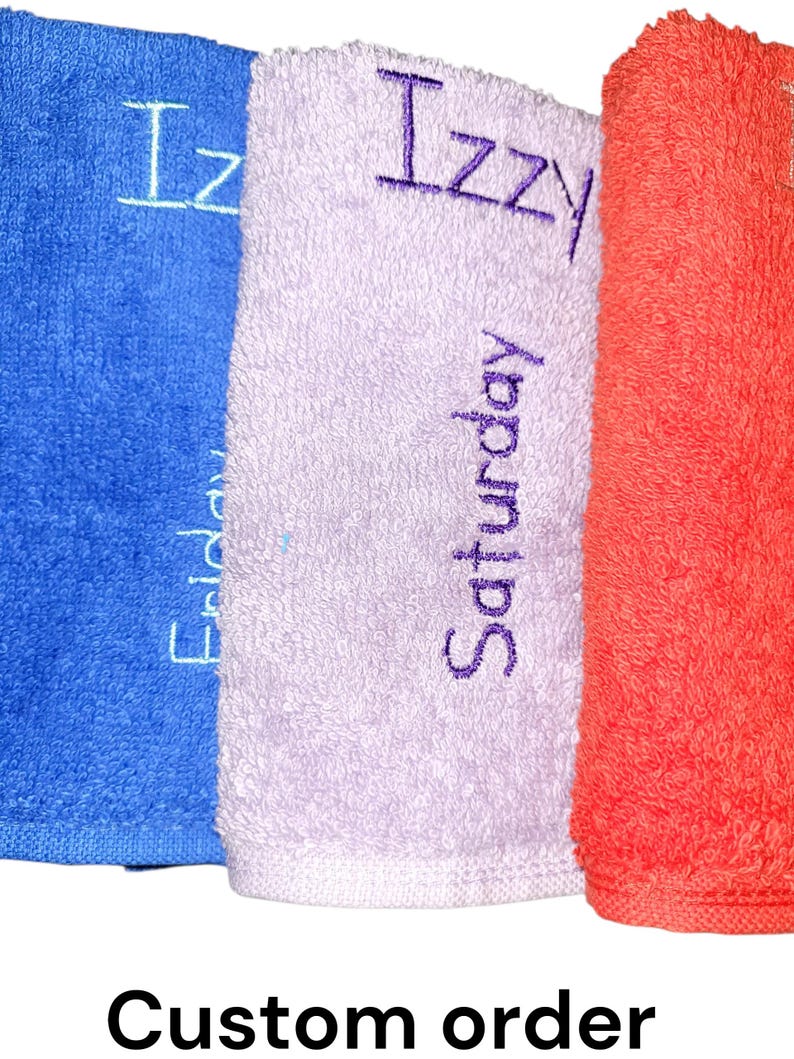 May include: Three folded terry cloth towels in blue, light purple, and orange. The blue towel features the embroidered name Izzy and the word Friday. The light purple towel displays the name Izzy and the word Saturday in dark purple thread.