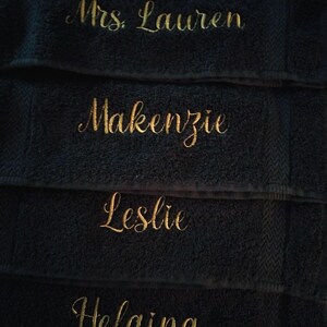 May include: A stack of four black towels, each featuring gold embroidered text. The names embroidered in a cursive script are Mrs. Lauren, Makenzie, Leslie, and Helaina, arranged from top to bottom on the textured fabric of the towels.