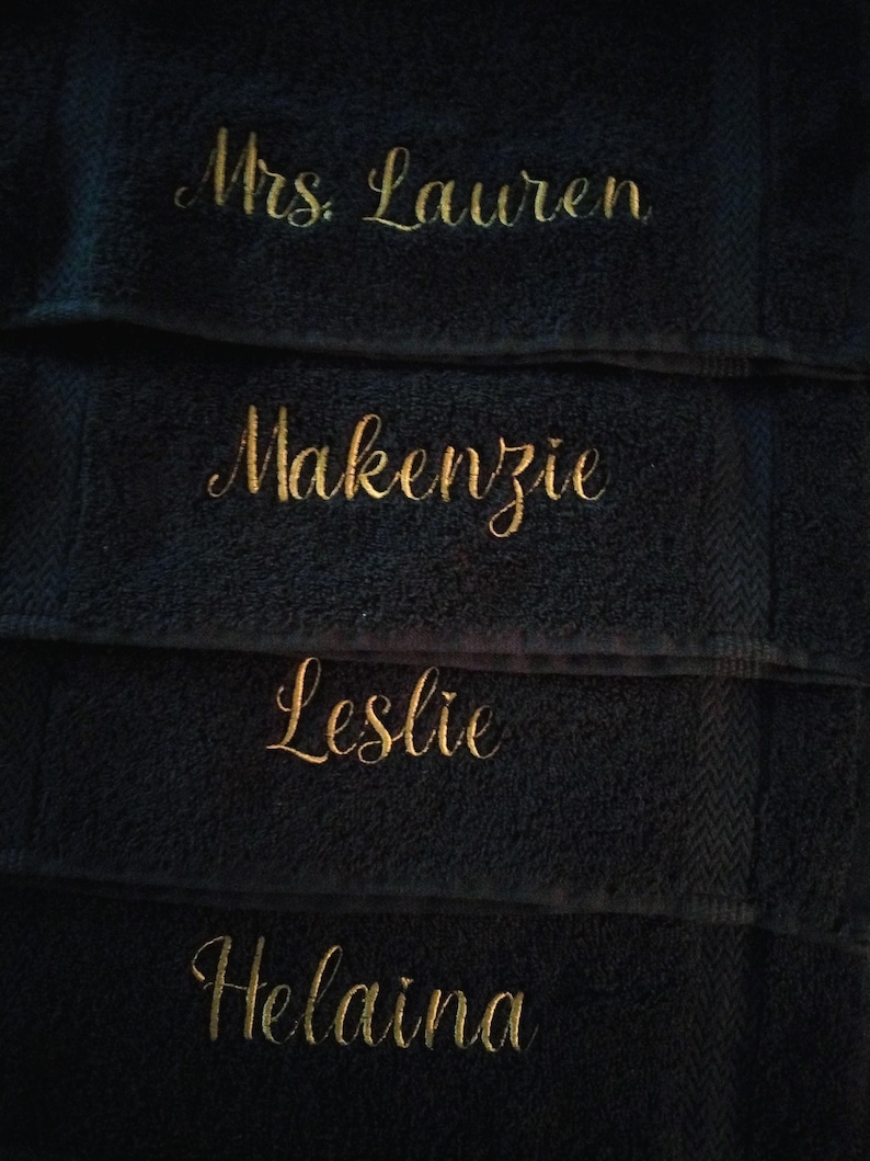 May include: A stack of four black towels, each featuring gold embroidered text. The names embroidered in a cursive script are Mrs. Lauren, Makenzie, Leslie, and Helaina, arranged from top to bottom on the textured fabric of the towels.
