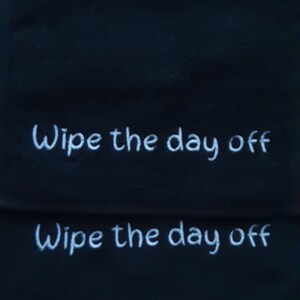 May include: A black fabric cloth featuring the white embroidered text "Wipe the day off" repeated twice in a horizontal orientation. The lettering is rendered in a simple, clean font style against the dark, solid-colored background material.