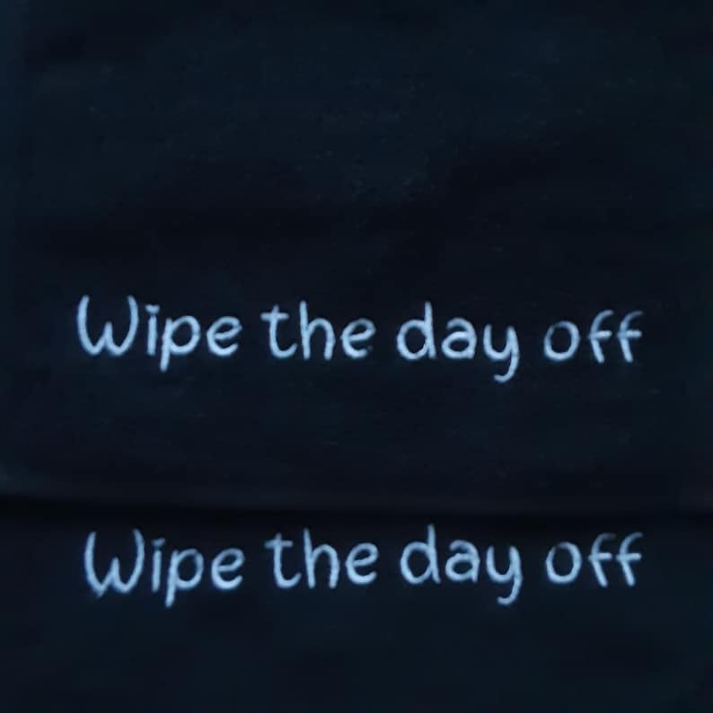 May include: A black fabric cloth featuring the white embroidered text "Wipe the day off" repeated twice in a horizontal orientation. The lettering is rendered in a simple, clean font style against the dark, solid-colored background material.