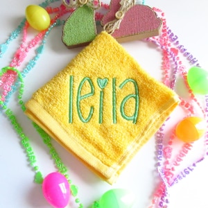 May include: A bright yellow square washcloth featuring the name leila embroidered in green thread with a small heart above the letter i. The washcloth is surrounded by colorful plastic Easter eggs and beaded necklaces with text.