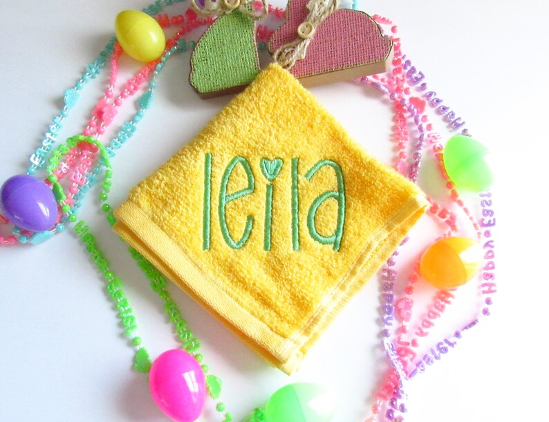 May include: A bright yellow square washcloth featuring the name leila embroidered in green thread with a small heart above the letter i. The washcloth is surrounded by colorful plastic Easter eggs and beaded necklaces with text.