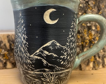May include: Ceramic mug with a hand-painted night scene. The design features a crescent moon, stars, mountains, and trees in white against a dark blue background. The rim and handle are a light teal color.