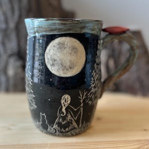 May include: A ceramic mug with a black background depicting a white full moon and a forest scene. The mug has a light blue rim and a handle with a red accent. The design includes a figure, a cat, and trees.