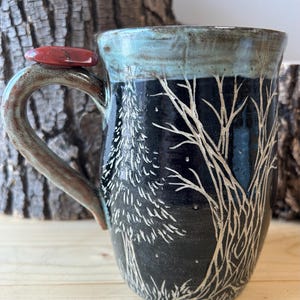 May include: A handmade ceramic mug with a black background and a white tree design. The rim is a teal color, and the handle is brown. A red stone is attached to the handle. The mug is a unique piece of art.
