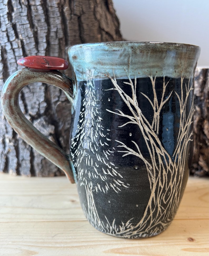 May include: A handmade ceramic mug with a black background and a white tree design. The rim is a teal color, and the handle is brown. A red stone is attached to the handle. The mug is a unique piece of art.