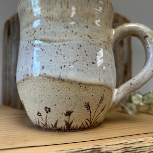 May include: A ceramic mug with a speckled cream and white glaze featuring a hand-drawn brown botanical illustration of wildflowers and grasses around the base. The mug has a rounded body, a wide rim, and a curved handle on the right side.