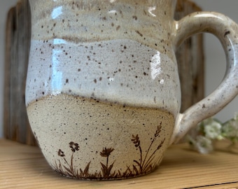 May include: A ceramic mug with a speckled cream and white glaze featuring a hand-drawn brown botanical illustration of wildflowers and grasses around the base. The mug has a rounded body, a wide rim, and a curved handle on the right side.