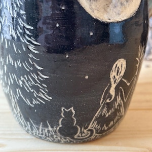 May include: Black ceramic mug with a white etched design of a full moon, stars, a cat, a person, and a tree. The mug has a dark, matte finish and a detailed, whimsical illustration.