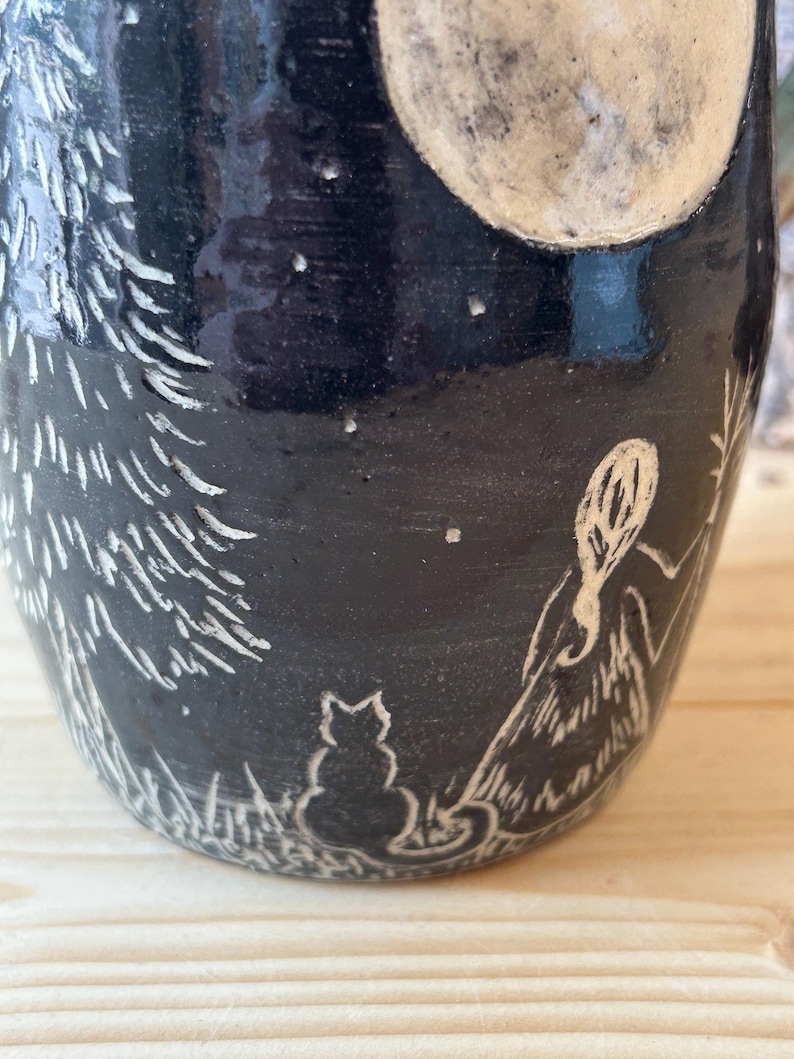 May include: Black ceramic mug with a white etched design of a full moon, stars, a cat, a person, and a tree. The mug has a dark, matte finish and a detailed, whimsical illustration.