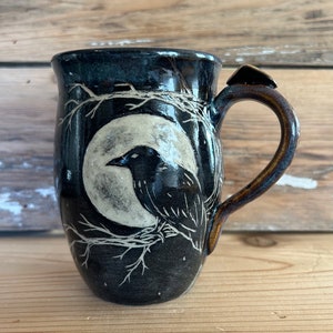 May include: A black ceramic mug featuring a raven and moon illustration. The raven sits on a branch against a full moon. The mug has a dark blue glaze, a brown handle, and a small black accent on the handle.