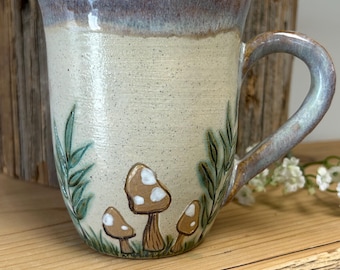 May include: A ceramic mug with a speckled cream-colored body, featuring a hand-painted design of three brown mushrooms with white spots and green leafy sprigs at the base. The rim and handle are finished in a glossy, variegated blue-grey glaze.