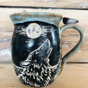 May include: A ceramic mug with a black design of a howling wolf under a full moon. The mug has a light blue rim and a decorative bird on the handle. The mug is on a wooden surface.